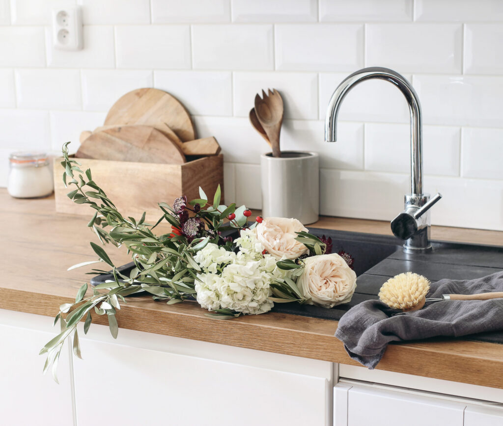 Closeup of kitchen interior. White brick wall, metro tiles, wooden countertops with kitchen utensils. Roses flowers in black sink.